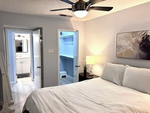 A hotel room with a bed, a ceiling fan, a nightstand with a lamp, a painting on the wall, and a view into the bathroom.