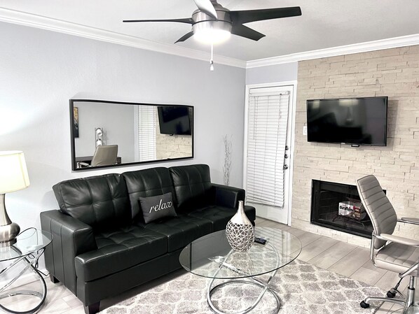 A modern living room with a black leather sofa, a glass coffee table, a fireplace, a TV mounted on the wall, and a ceiling fan.