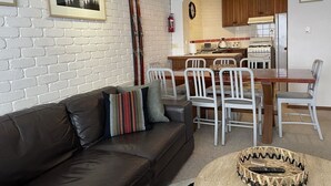 A living room with a brown leather sofa, a dining area with white chairs, a kitchen with wooden cabinets, and a white brick wall with a framed picture.