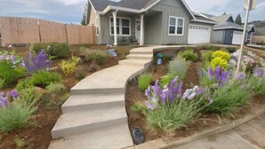 Una casa residencial con jardín delantero que incluye un camino de concreto, diversas plantas con flores y una cerca de madera.