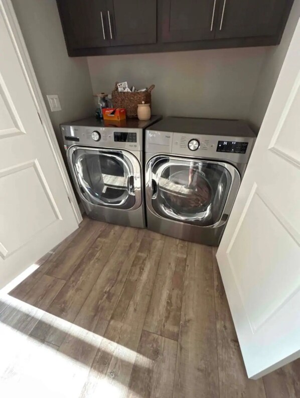 A laundry room with two stainless steel front-loading washers and dryers, wooden flooring, and dark cabinets above.