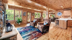 A wooden cabin interior with a stone fireplace, leather armchairs, a dining area, and a kitchen.