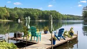 A wooden dock with colorful Adirondack chairs and umbrellas, overlooking a calm lake with a forested shoreline.