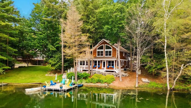 A two-story wooden house with a large porch, surrounded by trees and a lake.