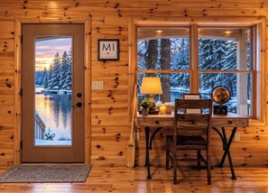 A wooden cabin interior with a door leading to a snowy landscape, a desk with a lamp, and a window with a view of snow-covered trees.