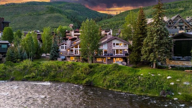 Uma pousada de montanha com vários edifícios, um rio ao primeiro plano e uma encosta arborizada.