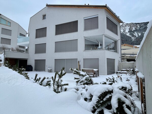 A modern multi-story building with large windows and balconies, surrounded by snow-covered ground and trees. - Family oasis with garden and fireplace (Lantsch/Lenz)