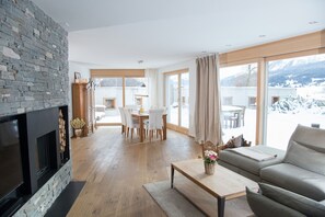 Ein modernes Wohnzimmer mit Kamin aus Stein, einer Couch, einem Essbereich mit Stühlen und großen Fenstern mit Blick auf die Berge.