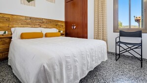 Een slaapkamer met een houten hoofdbord, een bed met witte beddengoed en mosterdgele kussens, een houten kledingkast, een stoel en een raam met uitzicht.