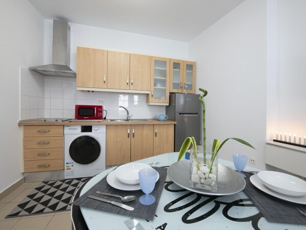 A modern kitchen with wooden cabinets, a stainless steel range hood, a red microwave, a washing machine, and a dining area with a table set for two.