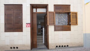 Une entrée d’immeuble avec une porte et des volets en bois, un intérieur visible comprenant une cuisine, et une enseigne avec un symbole en forme de « V ».