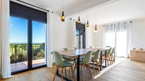 Un espace repas comprenant une grande table, des chaises et des luminaires suspendus. On peut admirer la verdure à travers les portes vitrées.