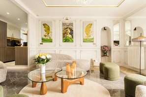 Una sala de estar moderna con una mesa de centro de vidrio, un sofá y un reposapiés verde. Hay tres obras de arte enmarcadas en la pared, una araña y una cocina al fondo.