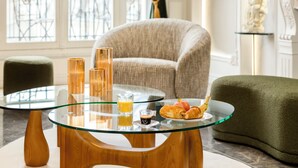 A glass-top table with drinks and pastries, a green armchair, and a beige sofa in a well-lit room with a view through a window. - Merveil Signature Suite Hoche - Eiffel Tower view (Paris)