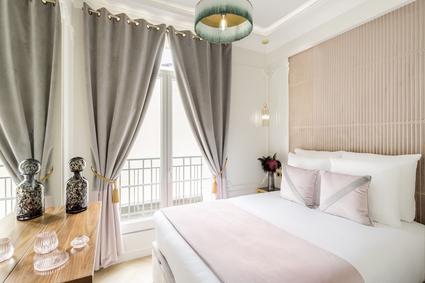 A bedroom with a large bed, grey curtains, a wooden nightstand with decorative items, and a view of the outside balcony.