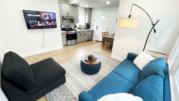 Una sala de estar moderna con un sofá azul, un sillón negro y una mesa de centro con una planta. Un televisor de pantalla plana está montado en la pared mostrando 'GODZILLA-KONG'.