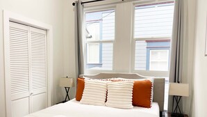 Una cama bien hecha con ropa de cama blanca y naranja, una pila de toallas blancas y una ventana con cortinas.