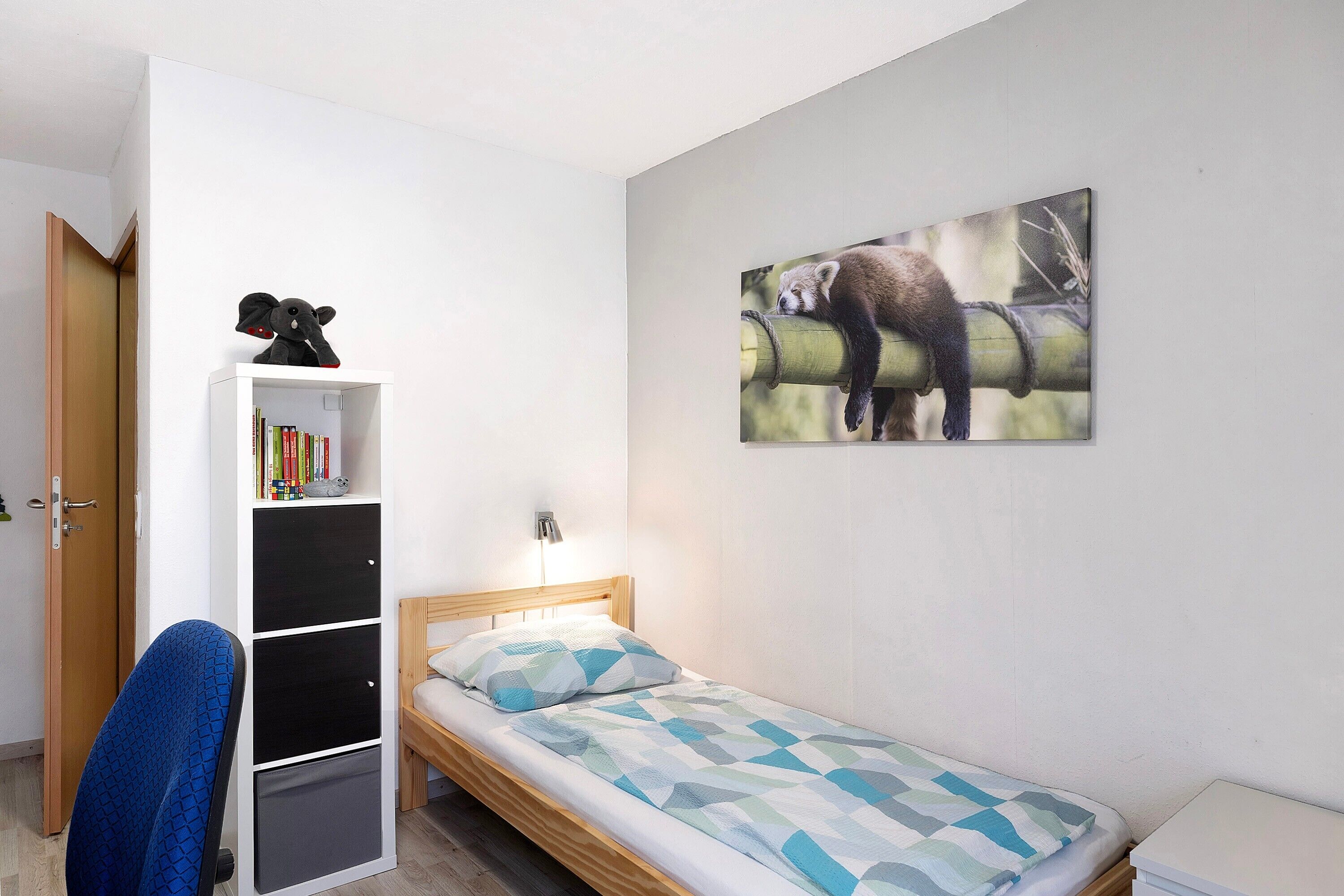 Une chambre avec un lit simple, un bureau avec une chaise, une bibliothèque et un tableau représentant un panda accroché au mur.