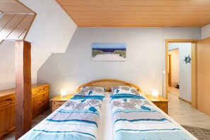 Quarto com cabeceira de madeira, duas camas com roupa de cama florida, abajures, um quadro de uma praia e um guarda-roupa de madeira.