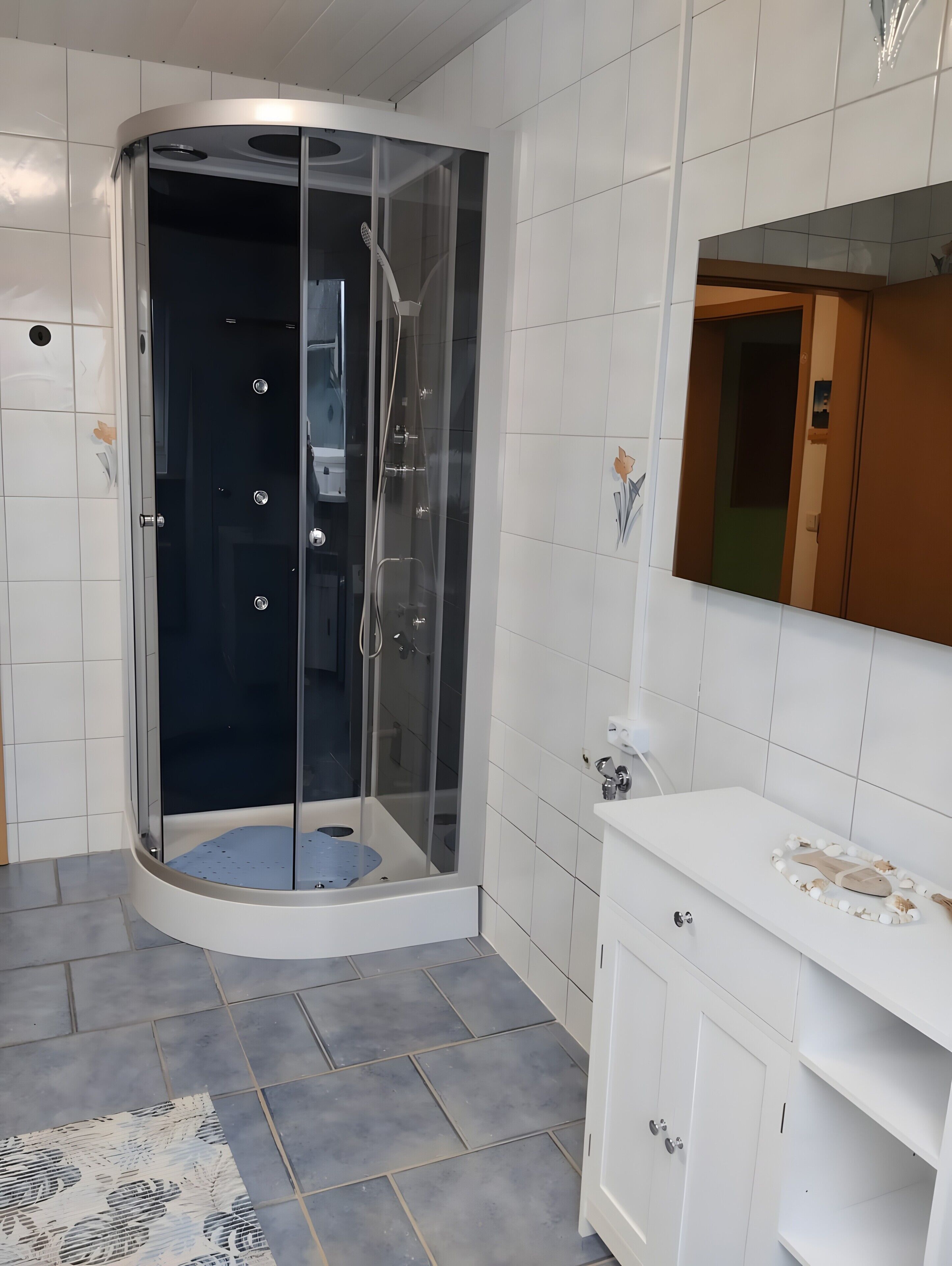 Een moderne badkamer met een douchecabine van glas, witte betegelde wanden, een wit aanrecht met een spiegel en een betegelde vloer.