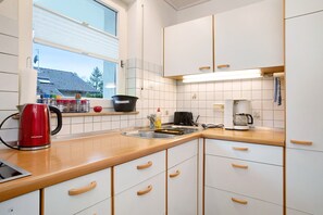 Une cuisine moderne avec des meubles blancs, un plan de travail en bois, une bouilloire rouge et une fenêtre donnant sur une maison.