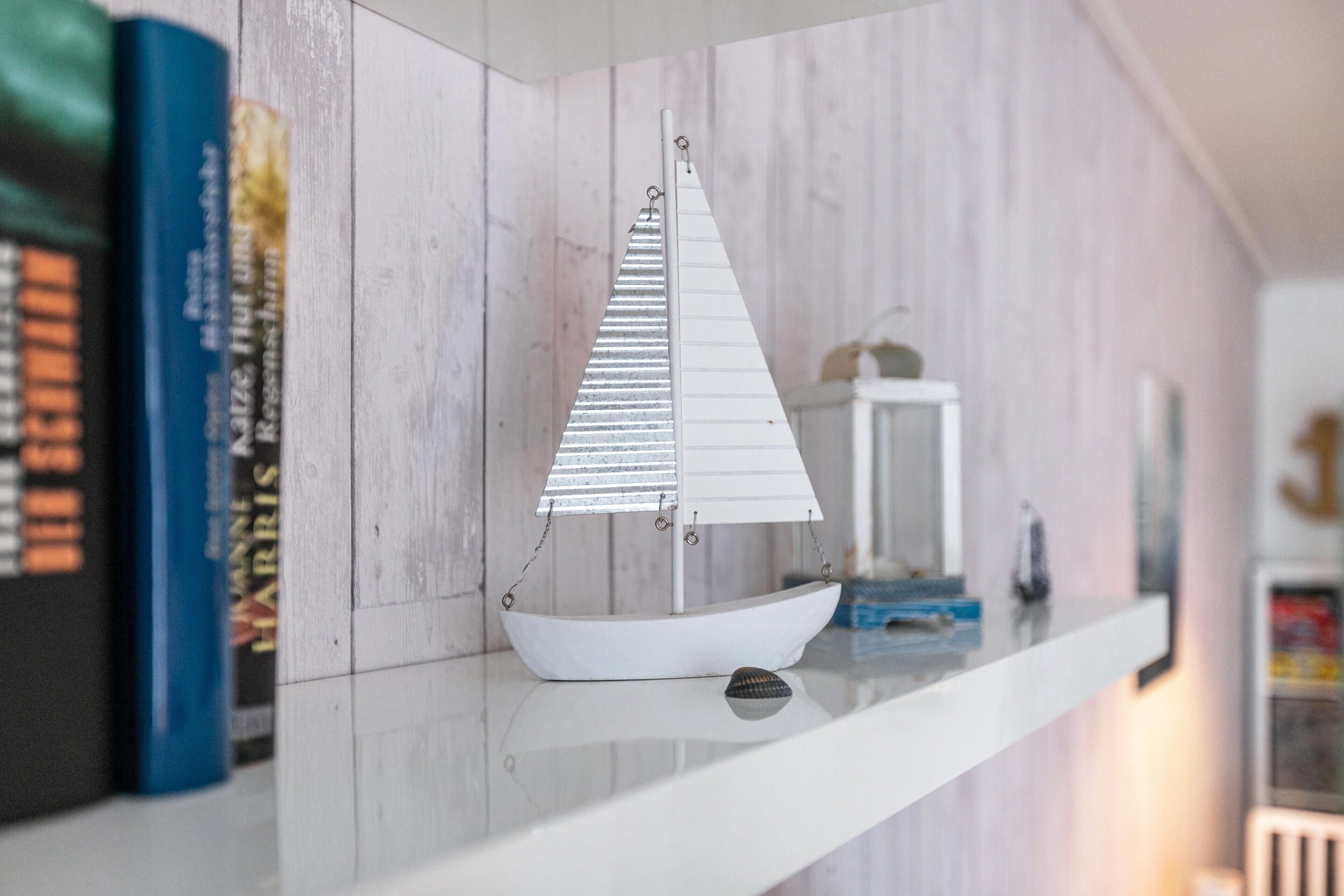 A model sailboat on a white shelf with books and a white lantern in the background.