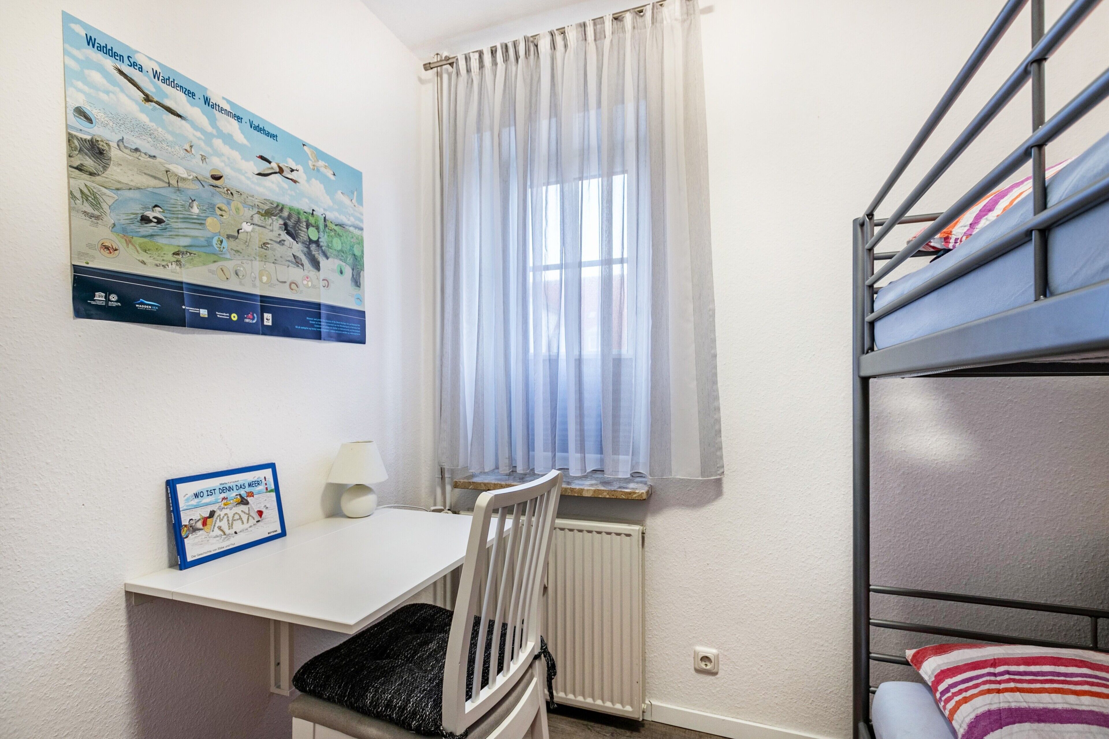 Ein Zimmer mit Etagenbetten, einem Schreibtisch, einem Stuhl, einem Heizkörper, einem fenster mit Vorhängen und einem Poster an der Wand.