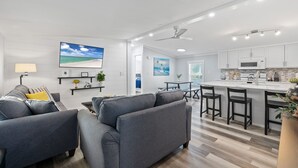 A modern living room with a gray sofa, a coffee table, and a TV. There is a kitchen area with a white countertop and bar stools. - Completely remodeled 3BR/2BA,PET friendly,5min from Hurlburt Field,fully fenced  (Mary Esther)