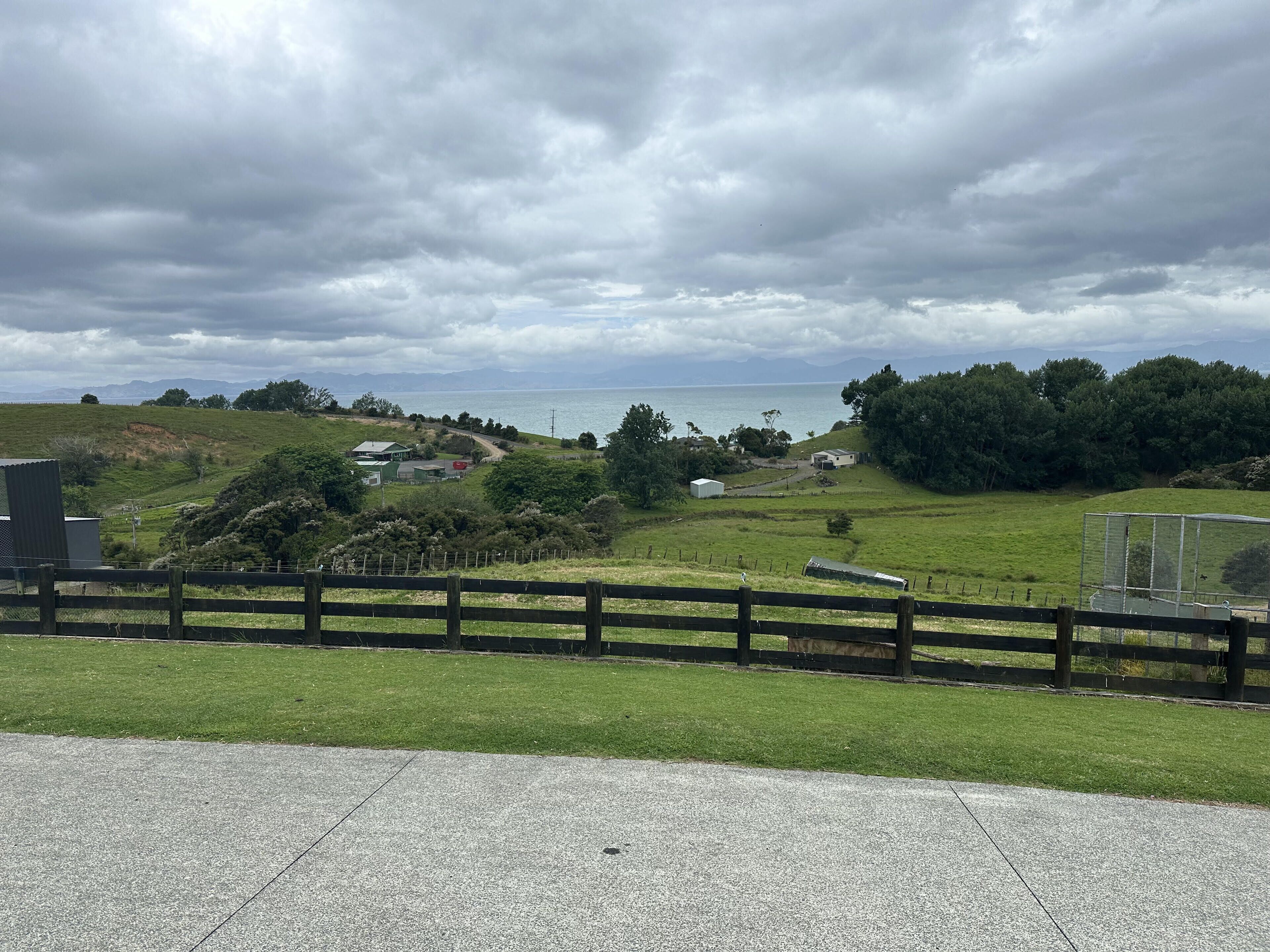 A coastal landscape with a grassy area, a wooden fence, and a paved path leading to a building.