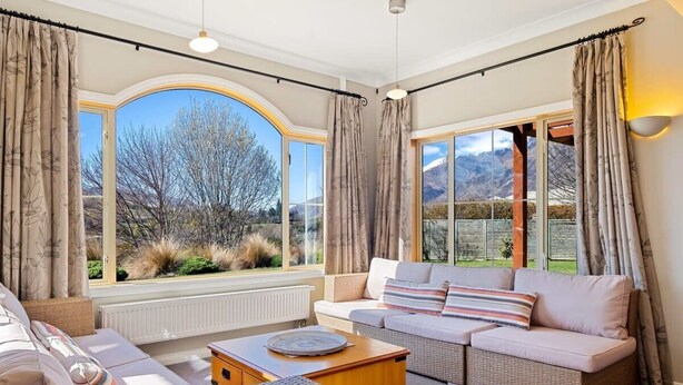 A cozy living room with a sofa, a coffee table, and a wicker chair. There are large windows with curtains, and a view of the outdoors.