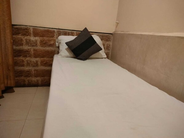A single bed with a white mattress and a dark striped pillow in a room with a brick wall and a plain white ceiling.