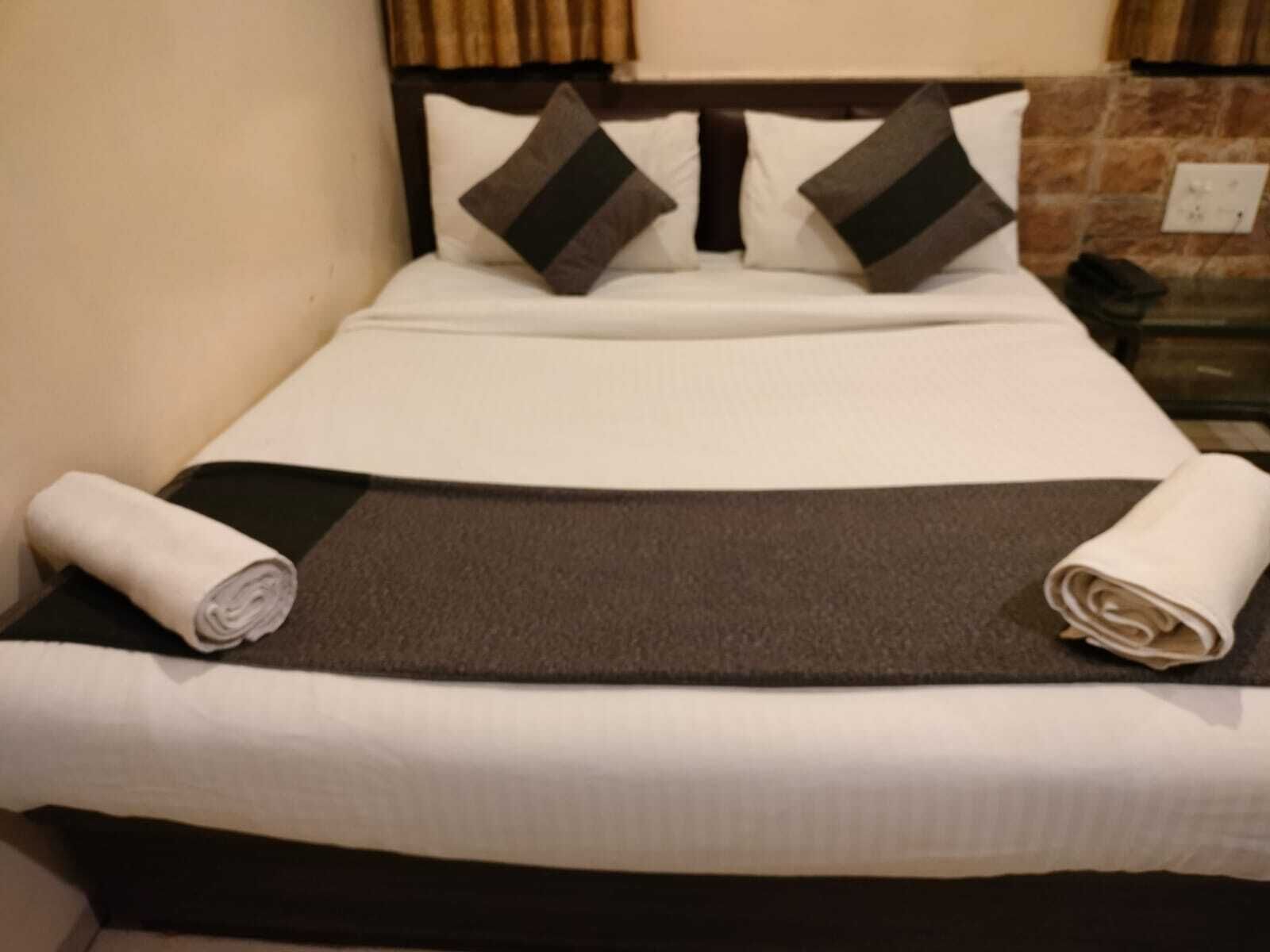 Cama bem arrumada com colcha branca e marrom, dois travesseiros e duas toalhas enroladas.