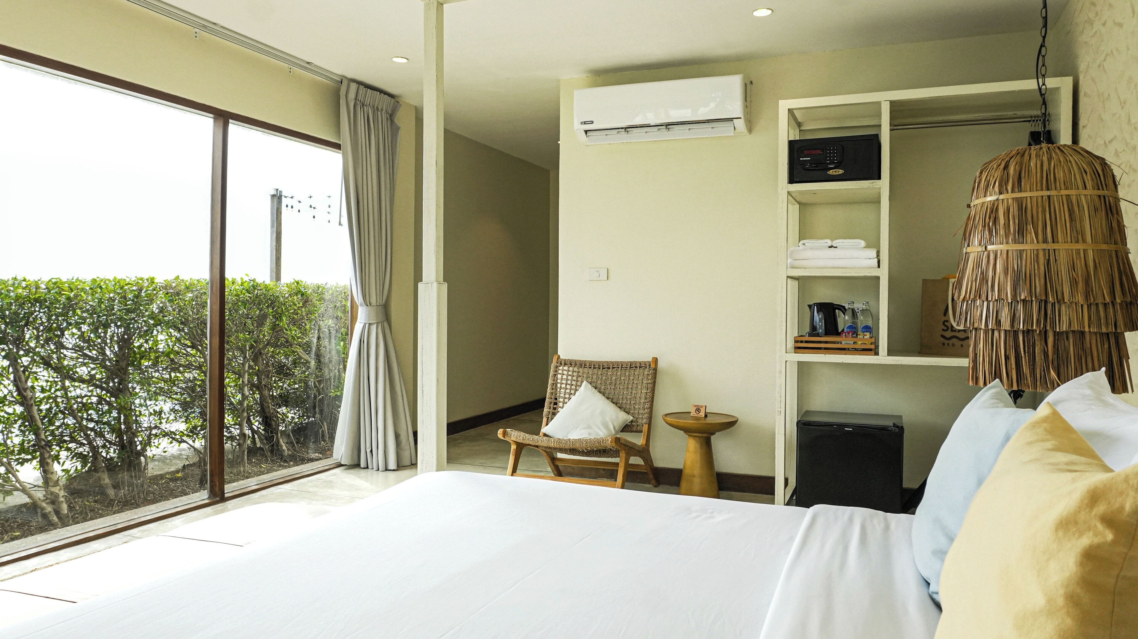 Ein Hotelzimmer mit einem großen Bett, einem Sessel, einem kleinen Tisch, einer Ablage mit Handtüchern und Pflegeprodukten sowie Blick auf Grünflächen.