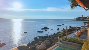 A coastal view with a pool area, rocky shoreline, and a distant island with a statue. - Sea Sun Bed & Beyond (Koh Samui)