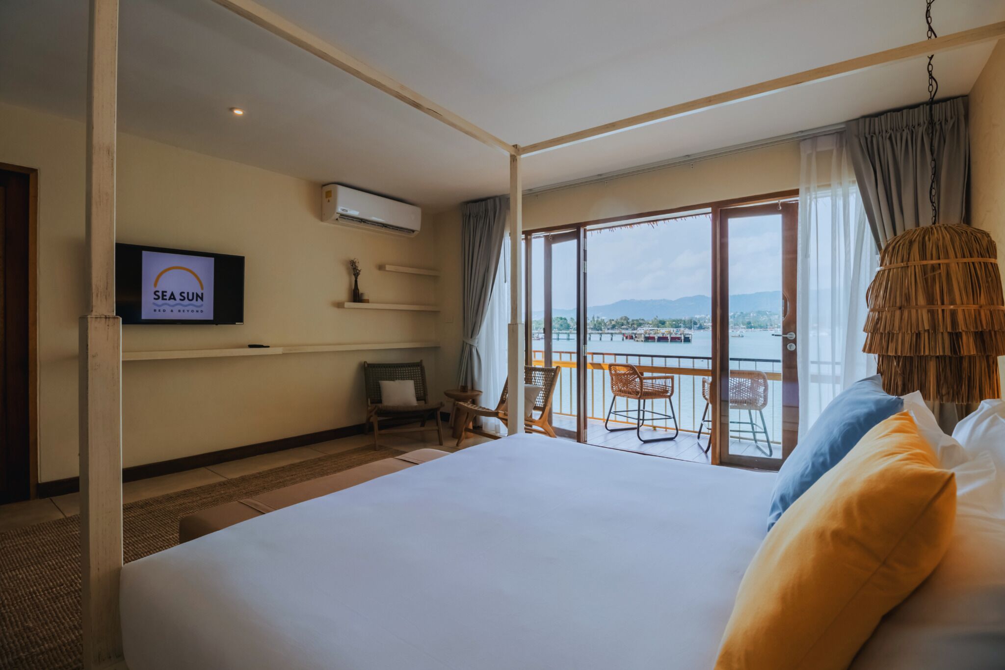 A hotel room with a large bed, a TV displaying 'SEA SUN', a balcony with chairs, and a view of the sea.