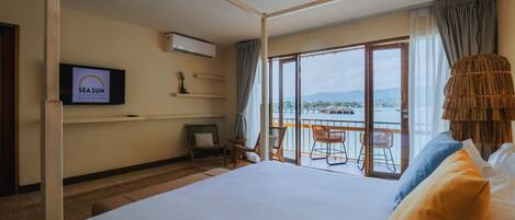 A hotel room with a large bed, a TV displaying 'SEA SUN', a balcony with chairs, and a view of the sea.
