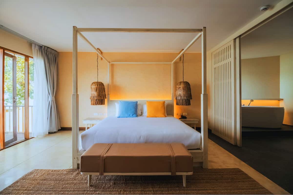 A four-poster bed with a white mattress, two pillows, a bedside table, a nightstand, a bench, a large window with curtains, a wall-mounted lamp, and a bathroom with a bathtub.