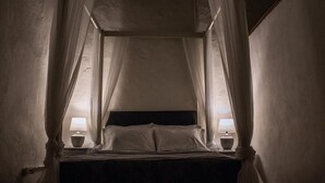 Ett svagt upplyst rum med en säng under ett himmelstält, två sänglampor och en enkel vägg.
