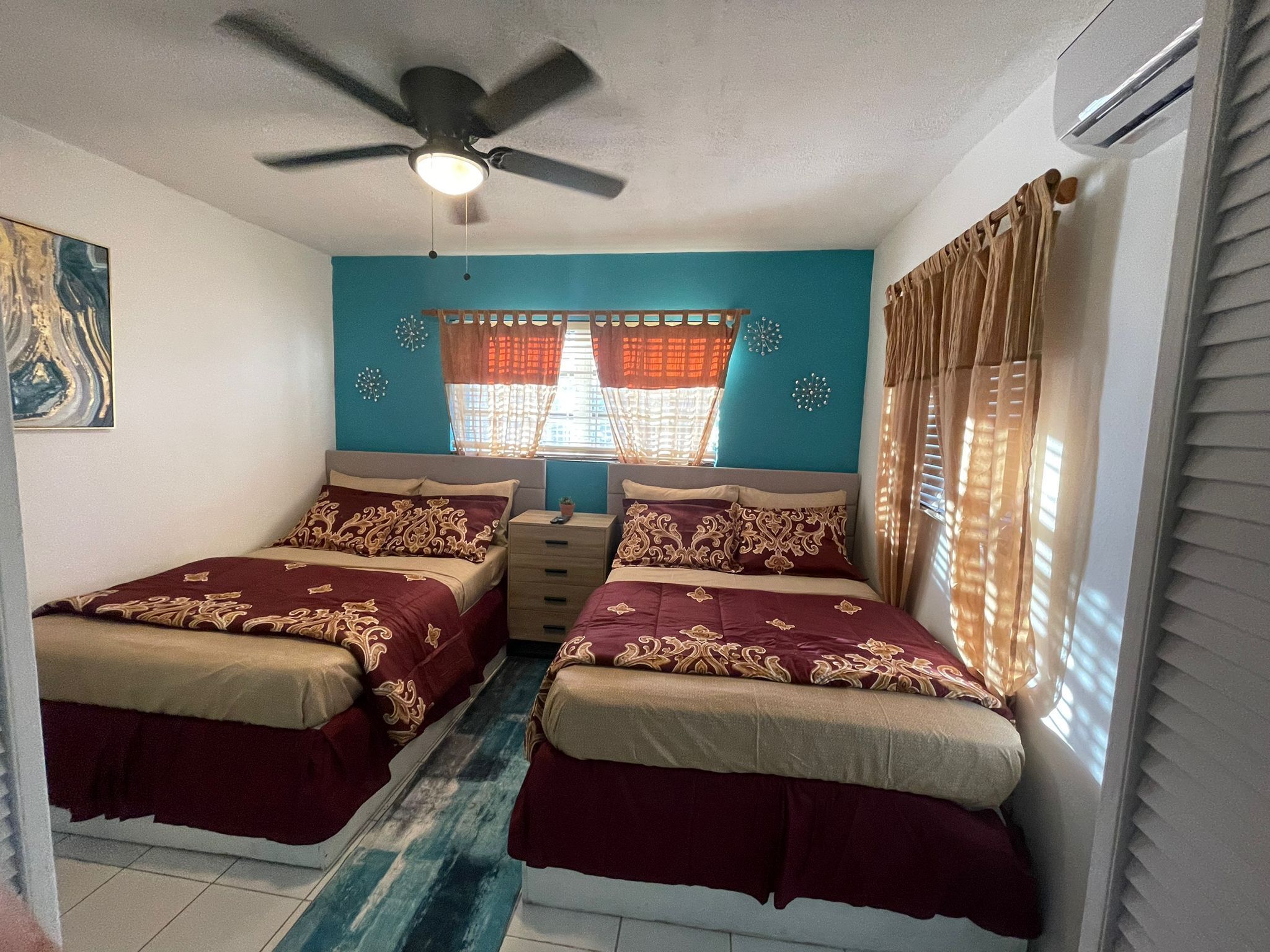 A bedroom with two beds, a ceiling fan, a window with curtains, and a small potted plant on a nightstand.