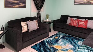 Un salon avec deux canapés noirs, un tapis aux motifs bleus et oranges, et un climatiseur fixé au mur.