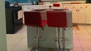Une cuisine avec des meubles blancs, un fourneau et un coin repas équipé de deux tabourets de bar rouges.