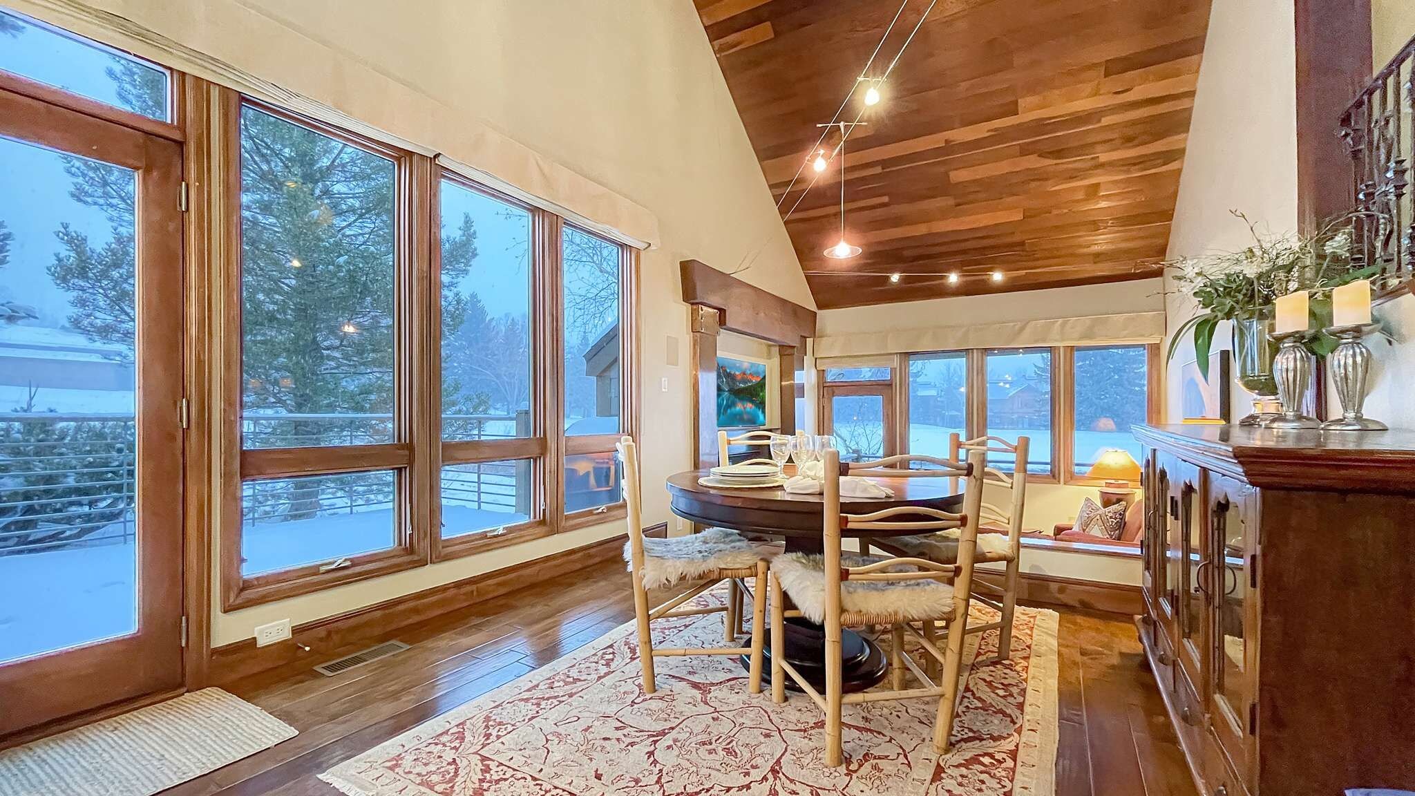 A dining area with a round table, chairs, and a view of a snowy landscape through large windows.