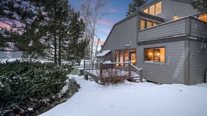 A modern two-story house with a deck, surrounded by snow-covered trees and a clear sky.