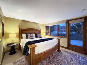 Een slaapkamer met een houten bed, een nachtkastje, een lamp en uitzicht op een besneeuwd landschap door een raam.