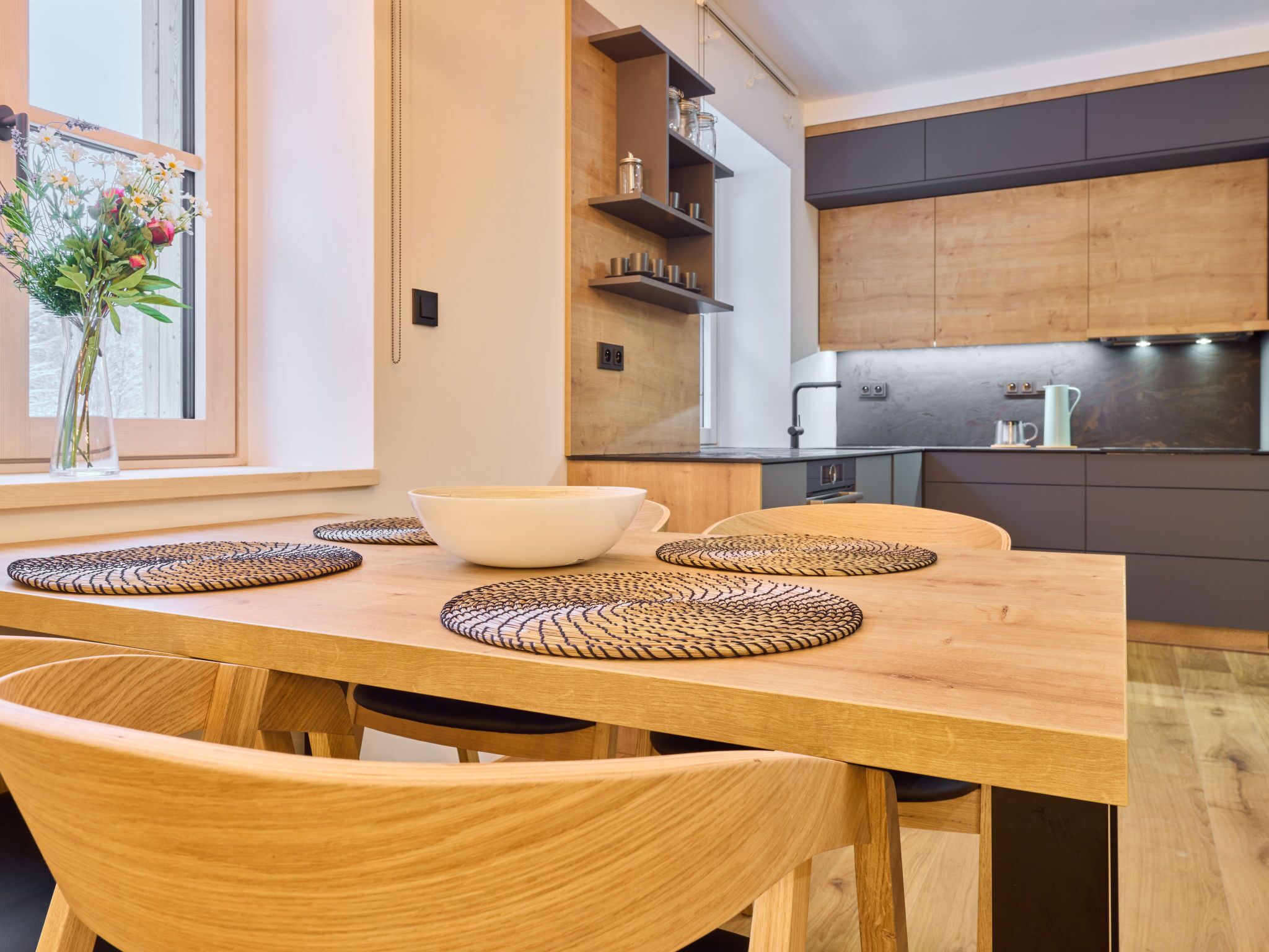 Una cocina moderna con armarios de madera, una mesa de comedor con individuales tejidos y un jarrón con flores en el alféizar.
