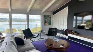 Ein modernes Wohnzimmer mit großem Fenster mit Blick auf ein Gewässer, einer Couch, zwei Sessel, einem Couchtisch und einem Flachbildfernseher.