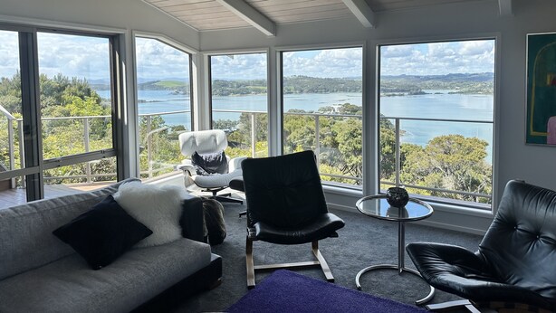 Ein geräumiges Zimmer mit großem Fenster und schöner Aussicht auf See und Hügel. Ein graues Sofa mit schwarzen Kissen, ein schwarzer Ledersessel und ein kleiner runder Tisch mit einer Dekoration sind vorhanden.