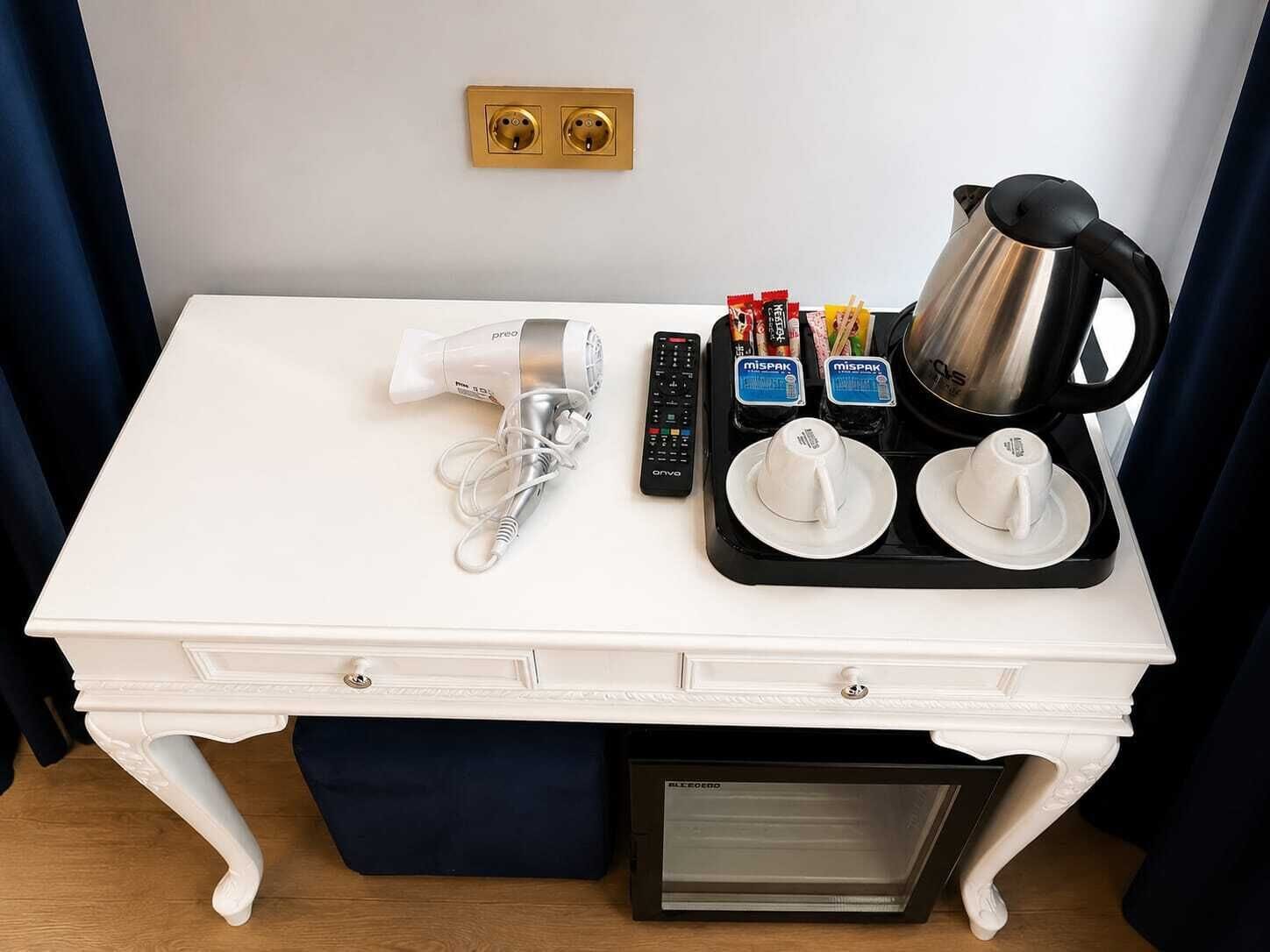 A white desk with a hairdryer, remote control, coffee maker, and tea packets.