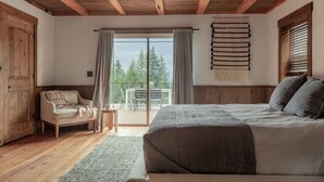 A bedroom with a large bed, wooden ceiling beams, a balcony with a view, and a rustic wooden door. - Ponderosa Glen @ Ashley Lake (Kila)