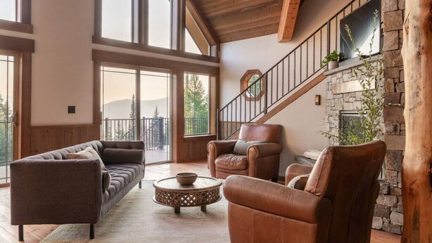 A spacious living room with a stone fireplace, leather armchairs, a sofa, and large windows offering a view of the outdoors. - Ponderosa Glen @ Ashley Lake (Kila)